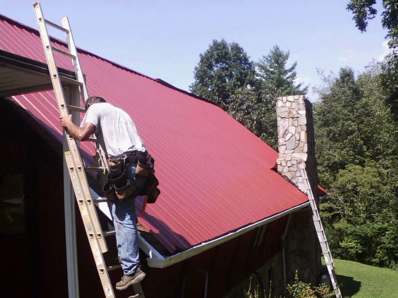 Roofer on a ladder beside a red metal roof and stone chimney — Soffit & Fascia Repair by Roofing Brothers in Savanna